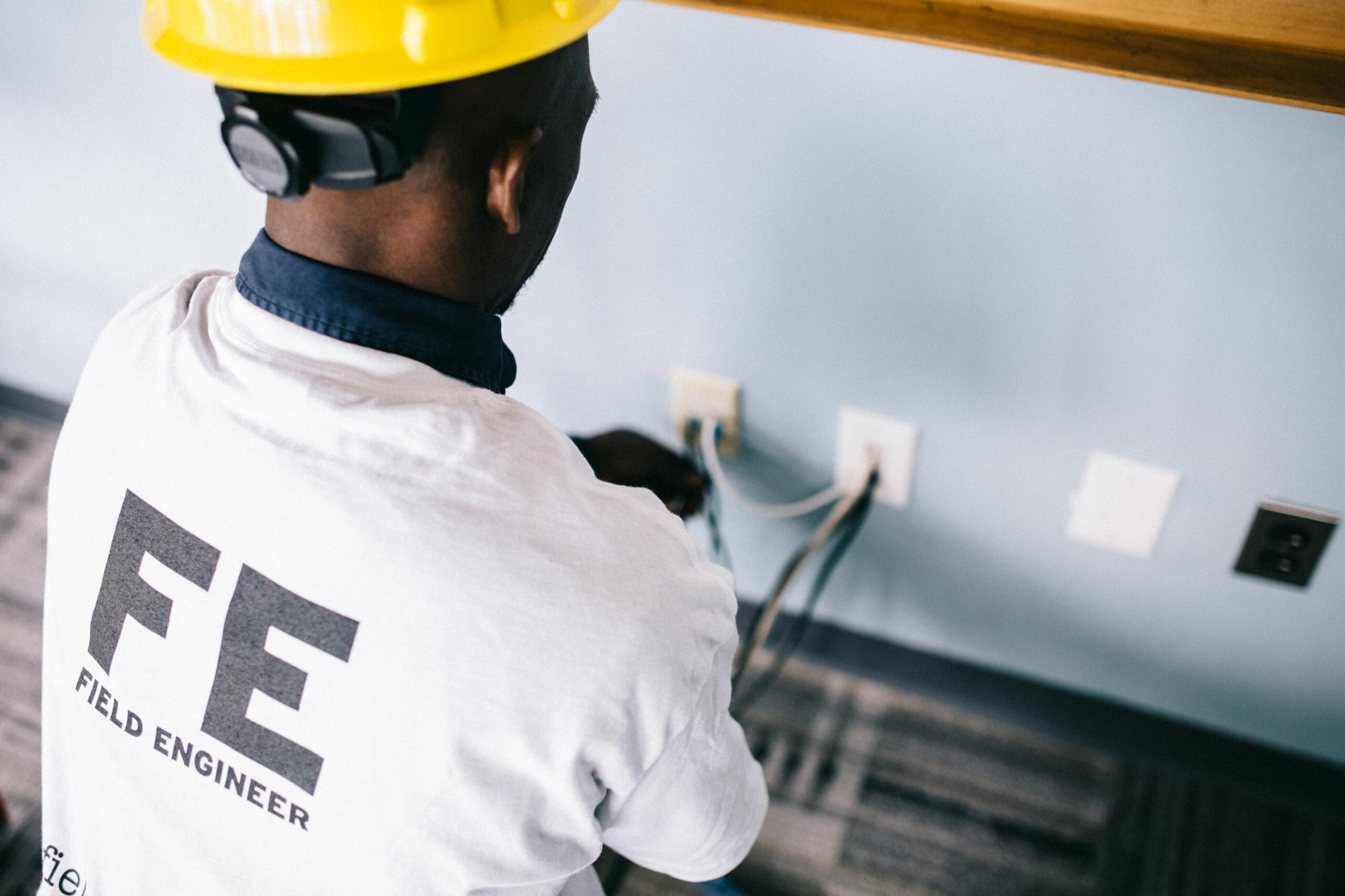 Home From above back view unrecognizable black field engineer wearing white shirt and protective hardhat sitting on floor and checking voltage in wall plugs